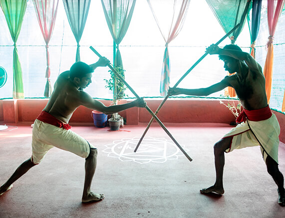 Two Kalaripayattu practitioners engaged in an intense wooden stick fight in a traditional training space